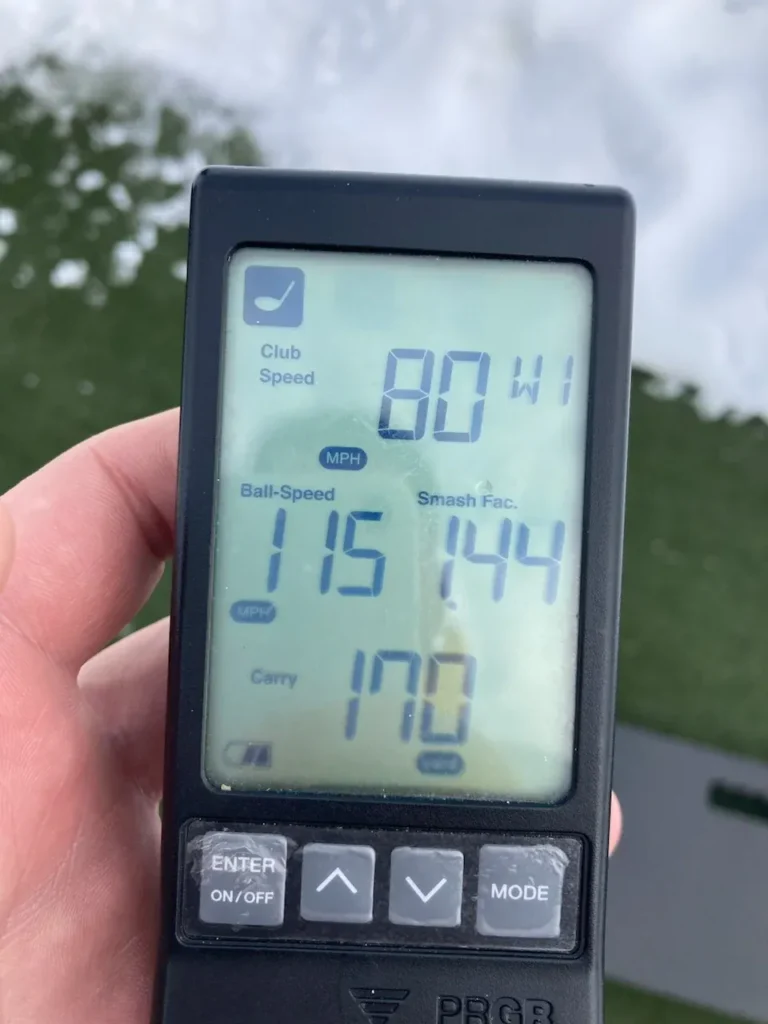 A hand holds a golf launch monitor displaying club speed of 80 mph, ball speed of 115 mph, a smash factor of 1.44, and a carry distance of 170 yards. A green surface and some water are in the background.