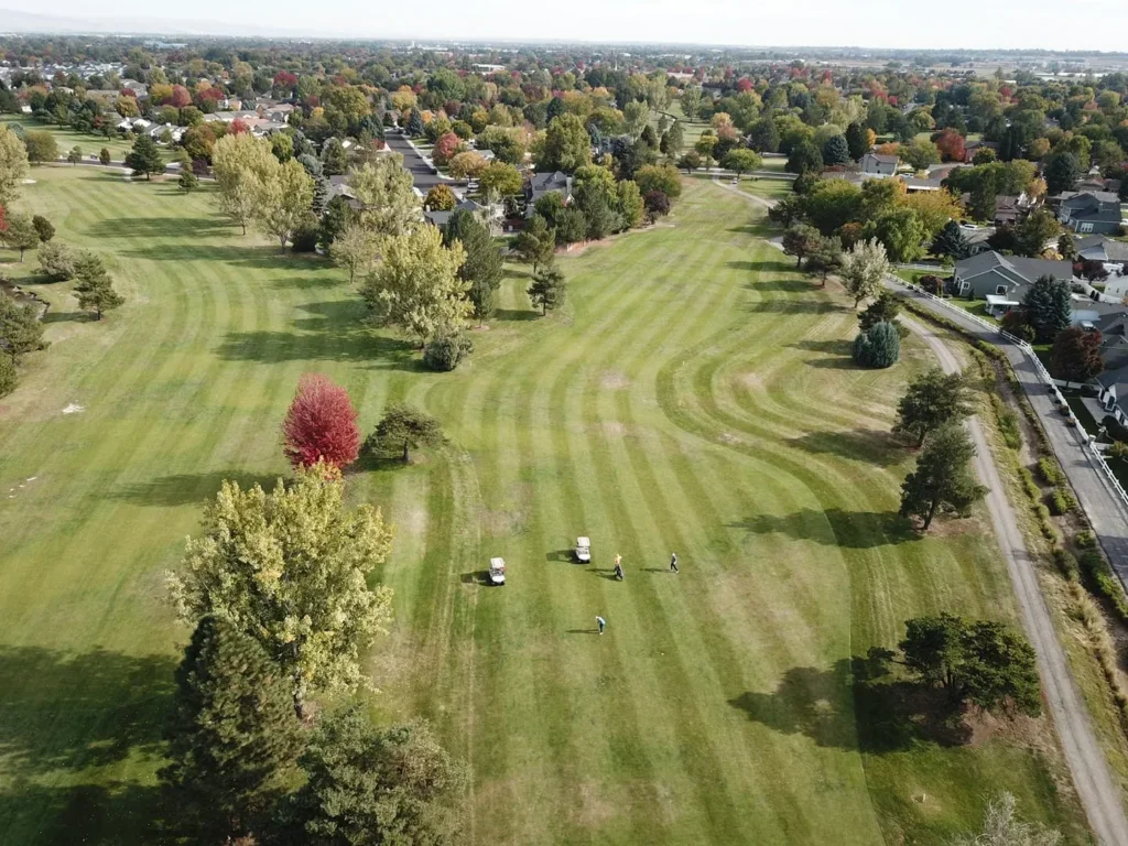 Aerial view of a golf course with green fairways, scattered trees, a few golf carts, and golfers. Surrounding the course are houses and streets, with autumn foliage visible throughout the landscape.