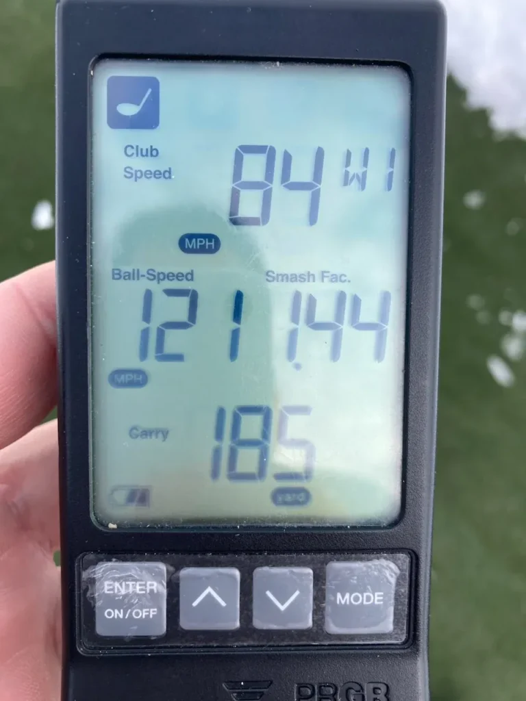 A hand holds a golf launch monitor displaying stats: club speed 84 mph, ball speed 121 mph, smash factor 1.44, and carry distance 185 yards. Snow or frost is visible on the ground in the background.