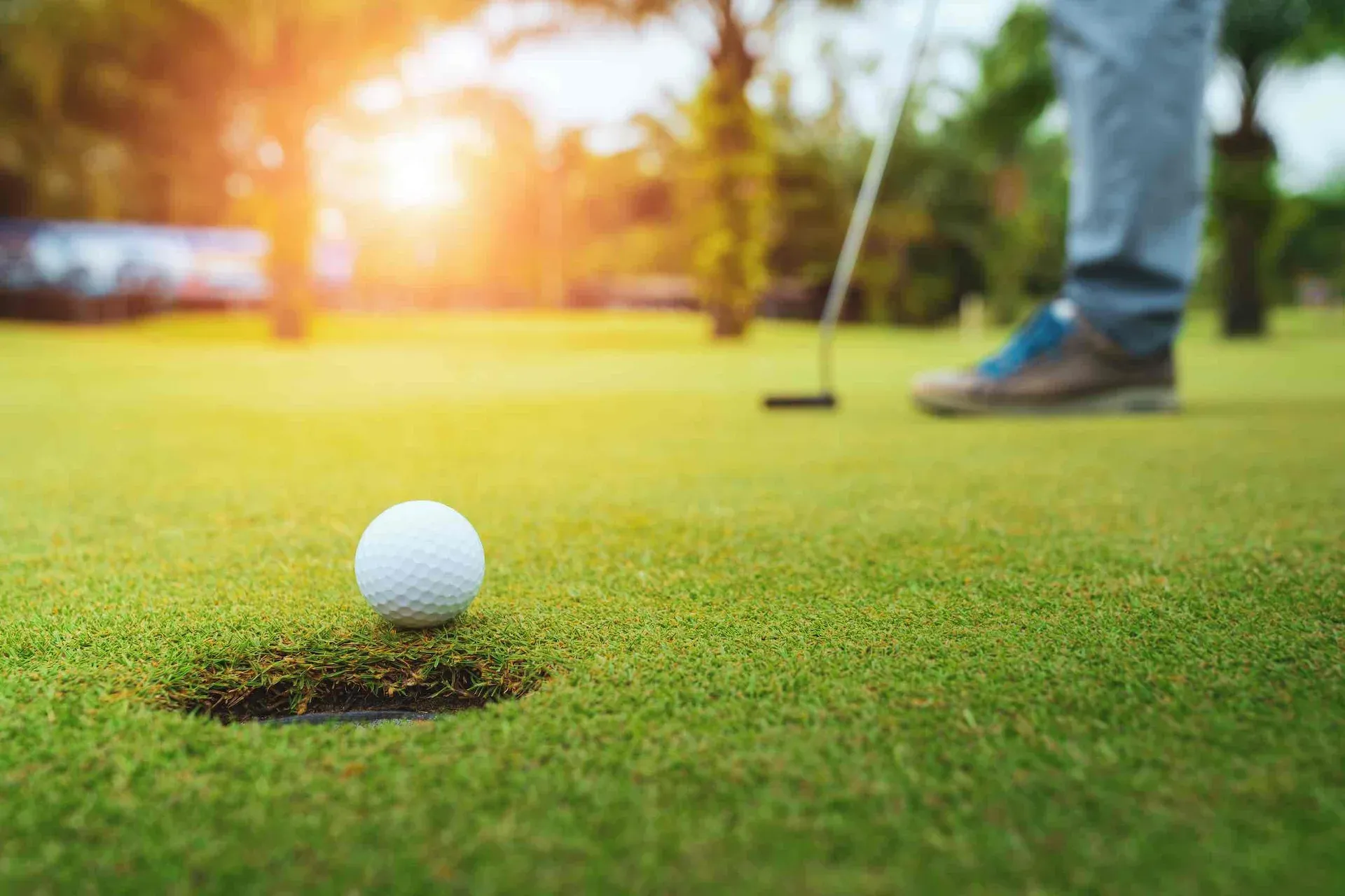 A close-up of a golf ball near a hole on a green, with a person standing in the background holding a golf club and sunlight shining through trees.