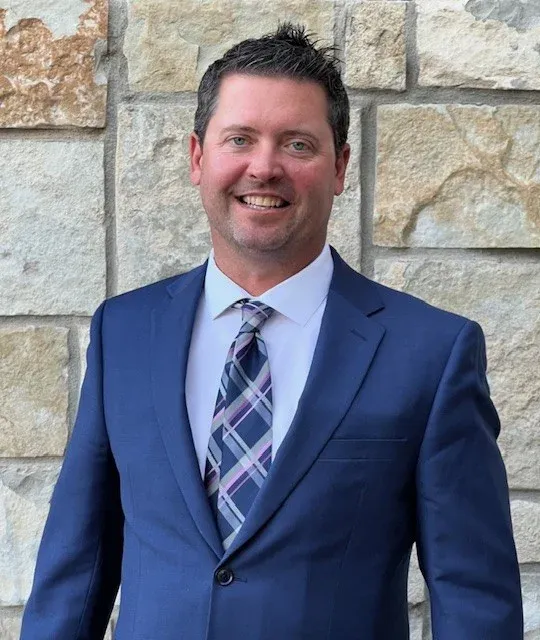 A man in a blue suit, white shirt, and plaid tie stands smiling in front of a stone wall.